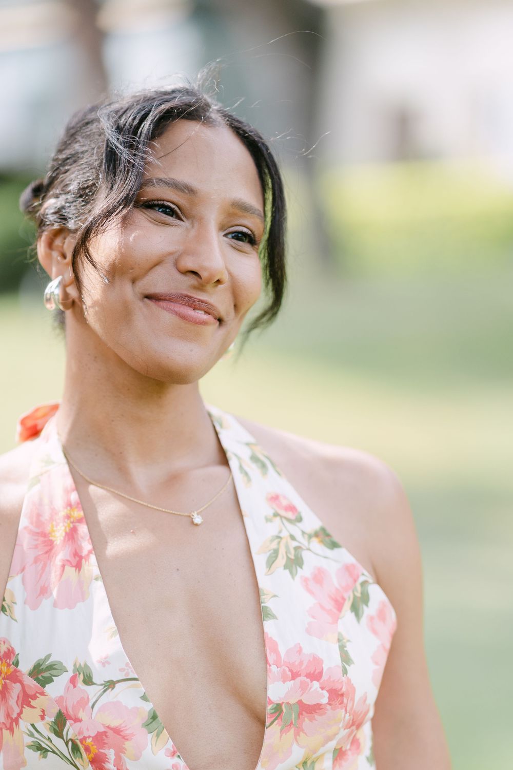 Woman in floral pink and white dress smiles joyfully while posing outdoors in natural lighting.