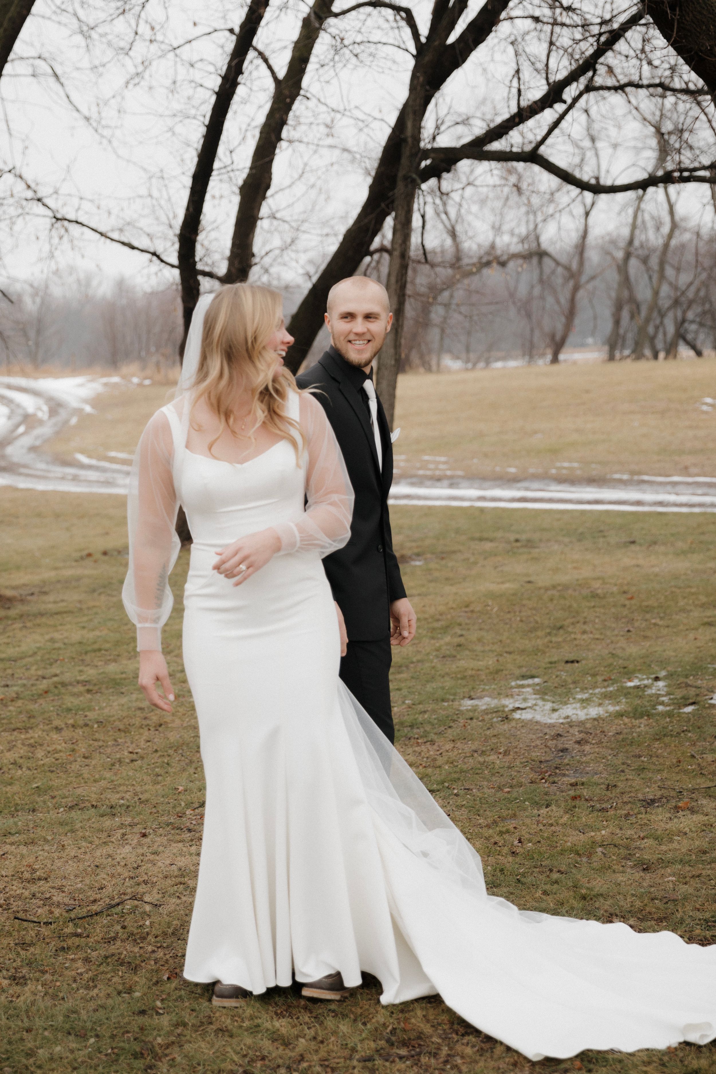Dancing couple in winter wedding attire twirls gracefully on snowy ground beneath bare tree branches.