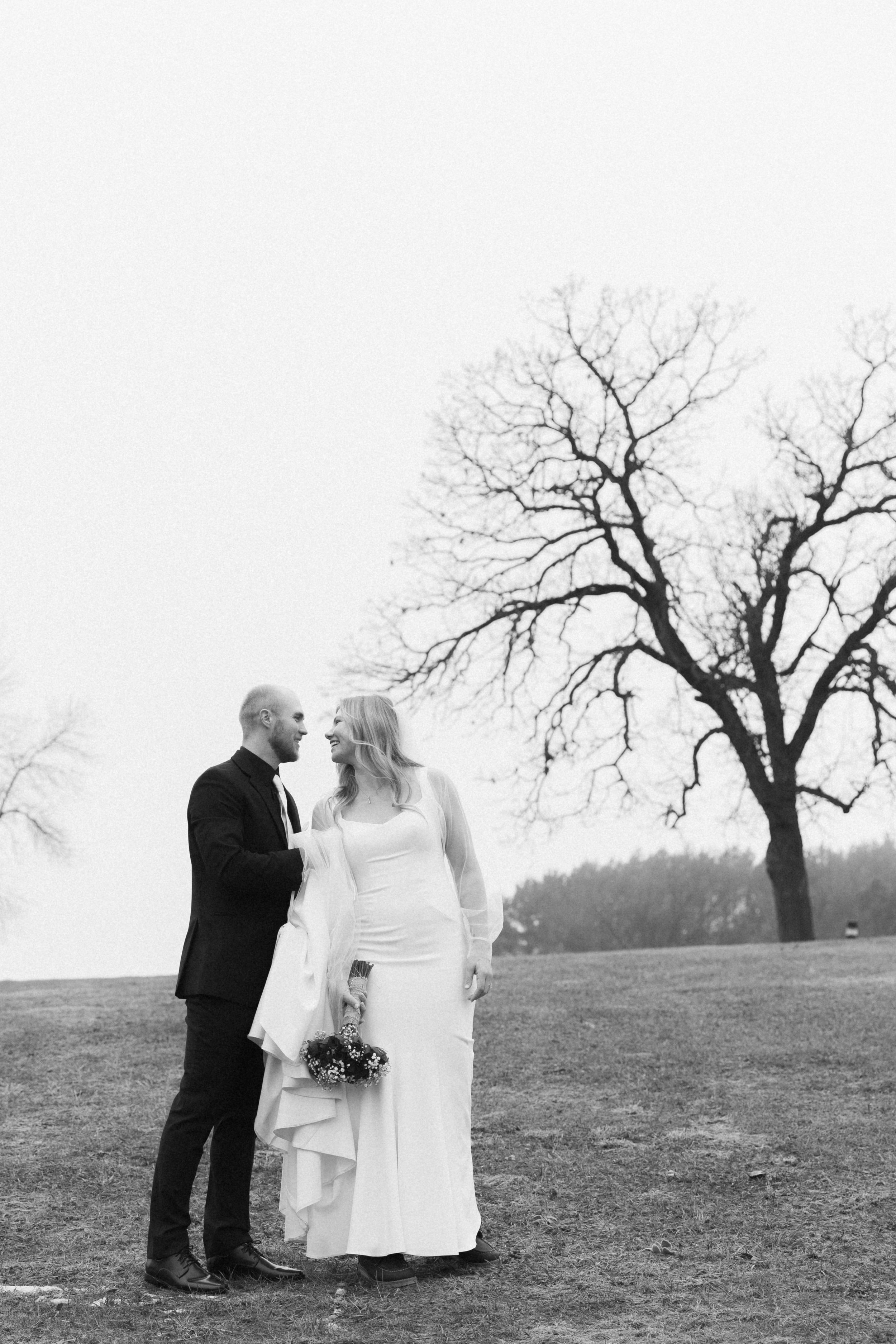 Black and white panoramic wedding photo showing a loving couple on a hilltop with bare winter trees in the background.