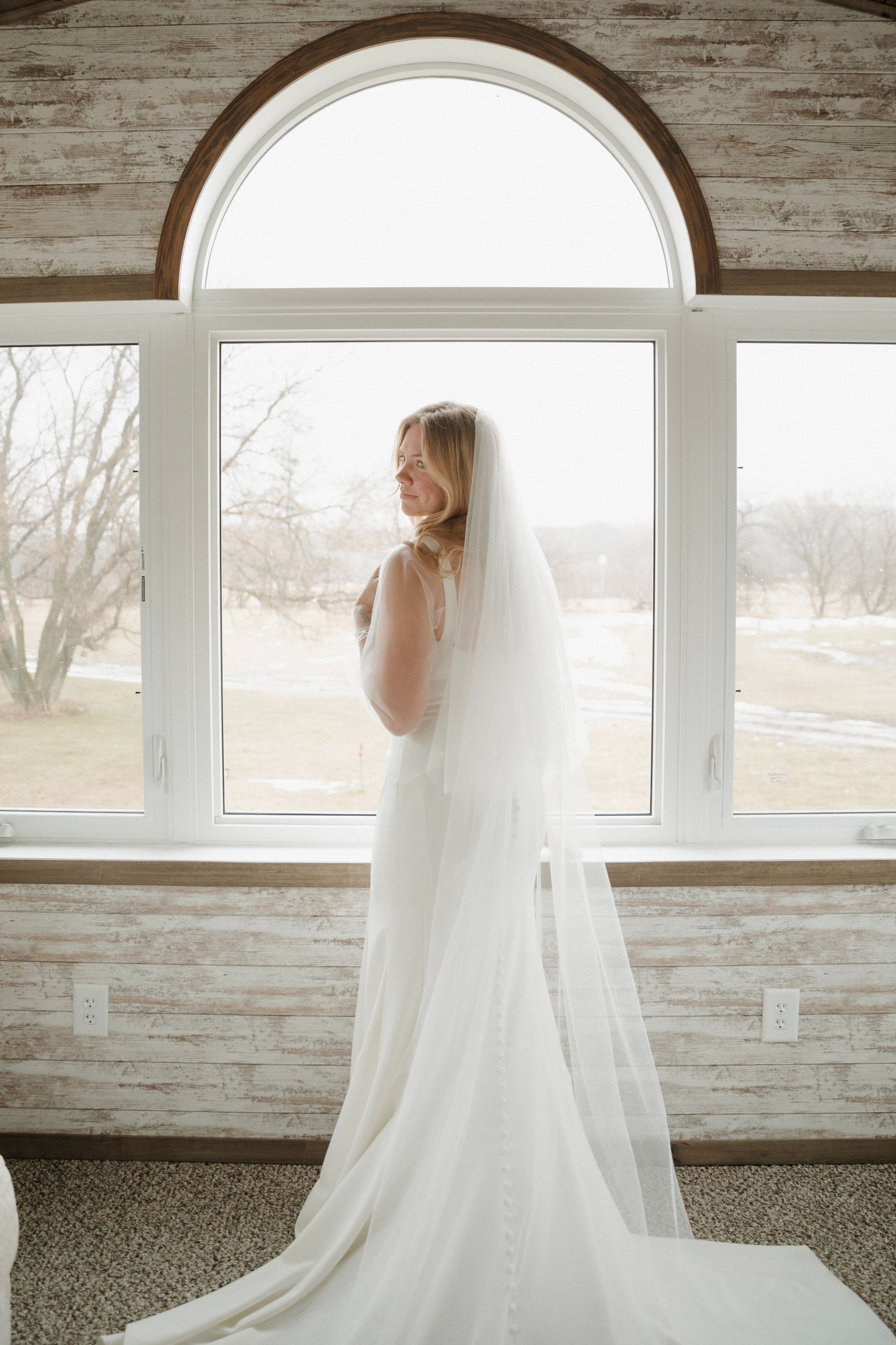 Elegant white wedding dress with flowing veil photographed in a rustic room with wooden walls and large windows.