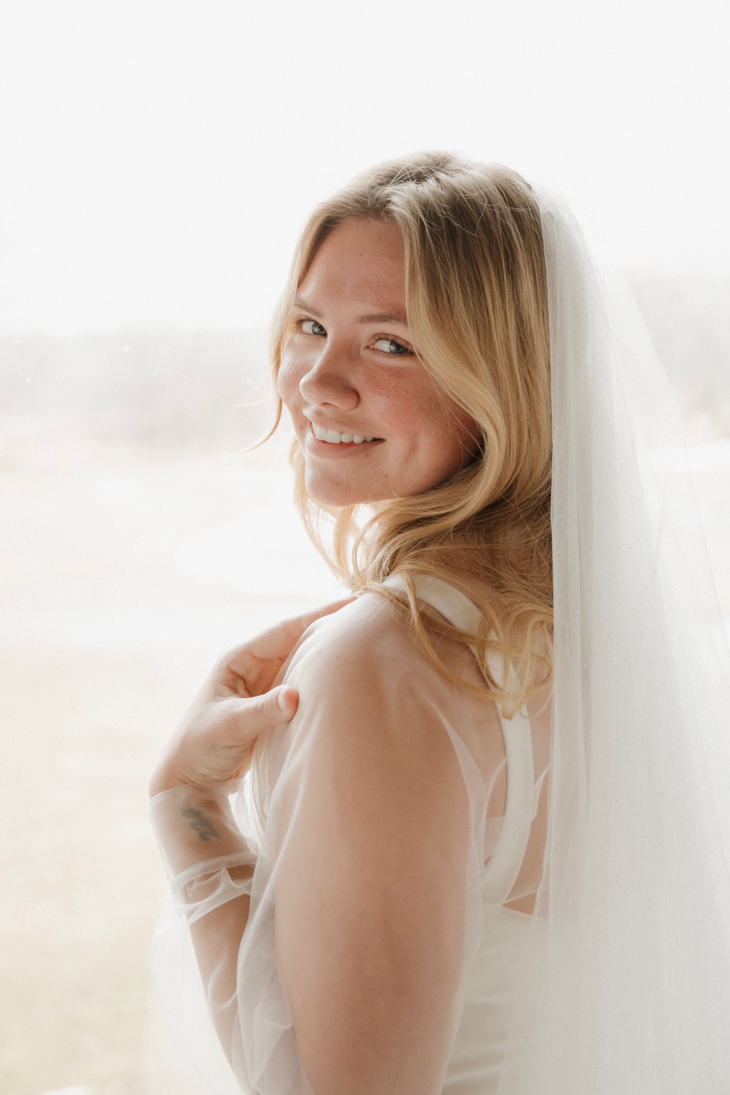 Bright and ethereal portrait of someone in white with blonde hair glancing over their shoulder and smiling.