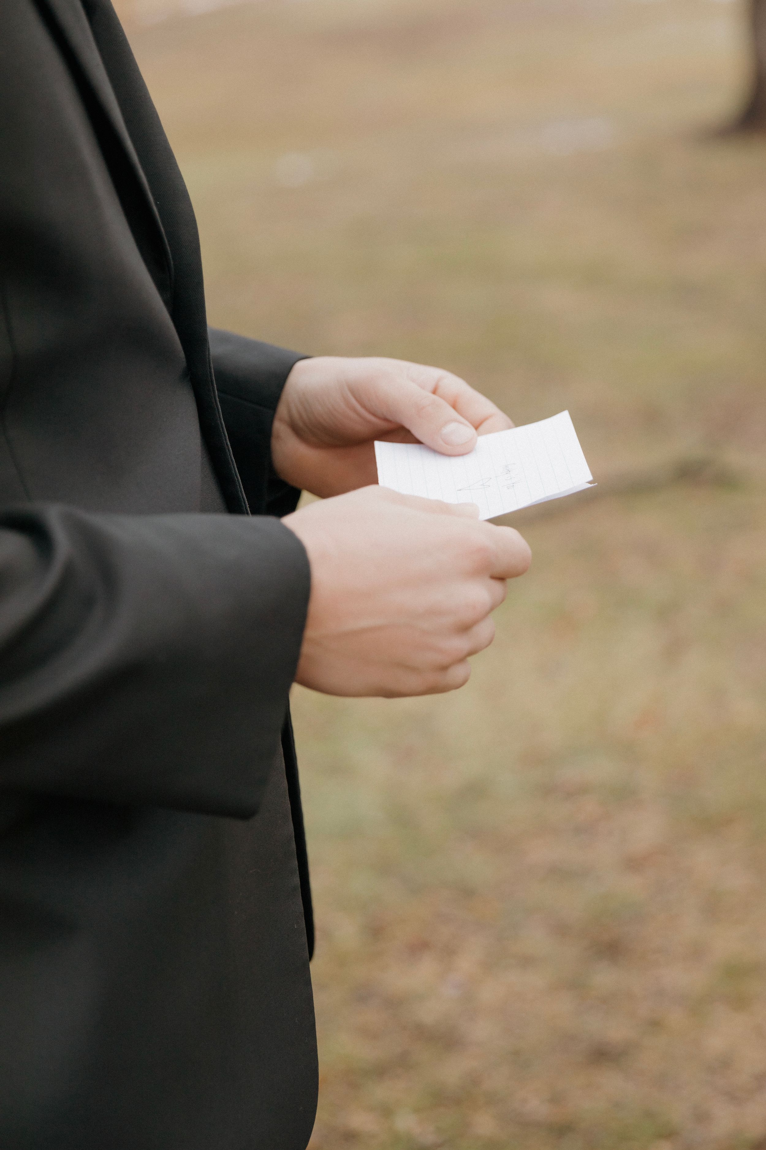 Hands in formal black attire holding a small white card or paper outdoors.