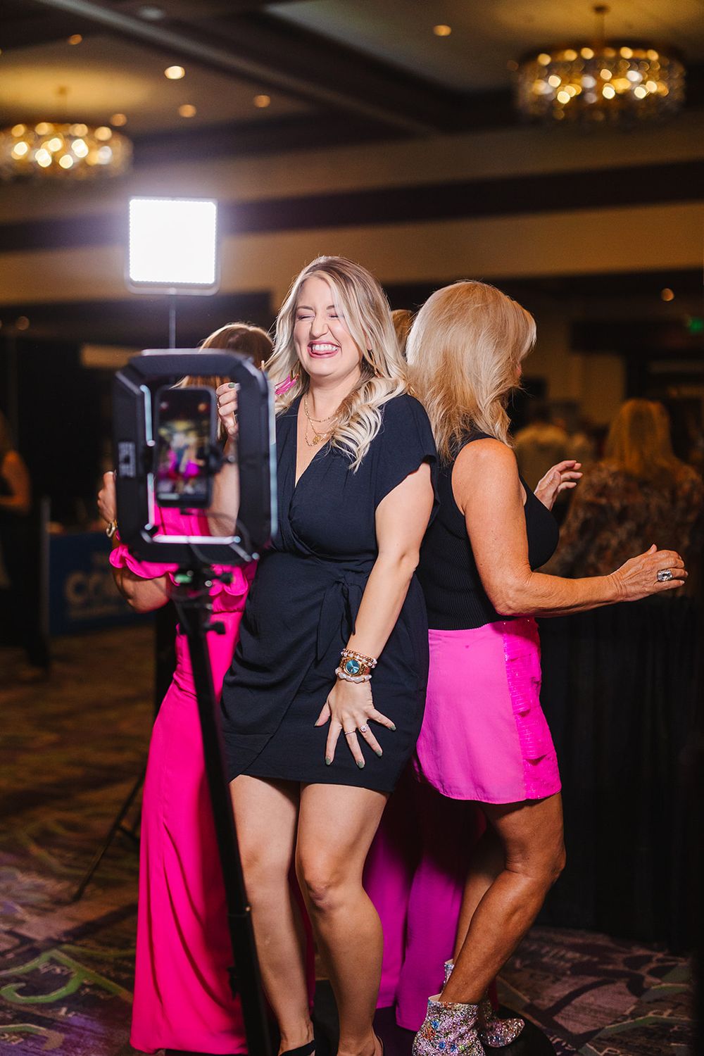 Dancing partygoers in black and hot pink dresses celebrate on a hotel ballroom dance floor under ambient lighting.
