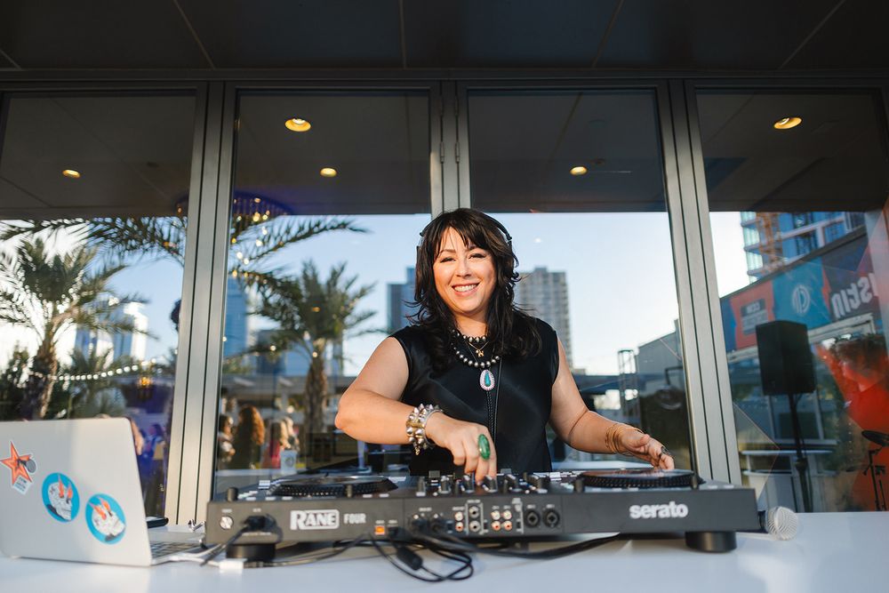DJ performs on turntables behind a storefront window with palm trees visible in the background.