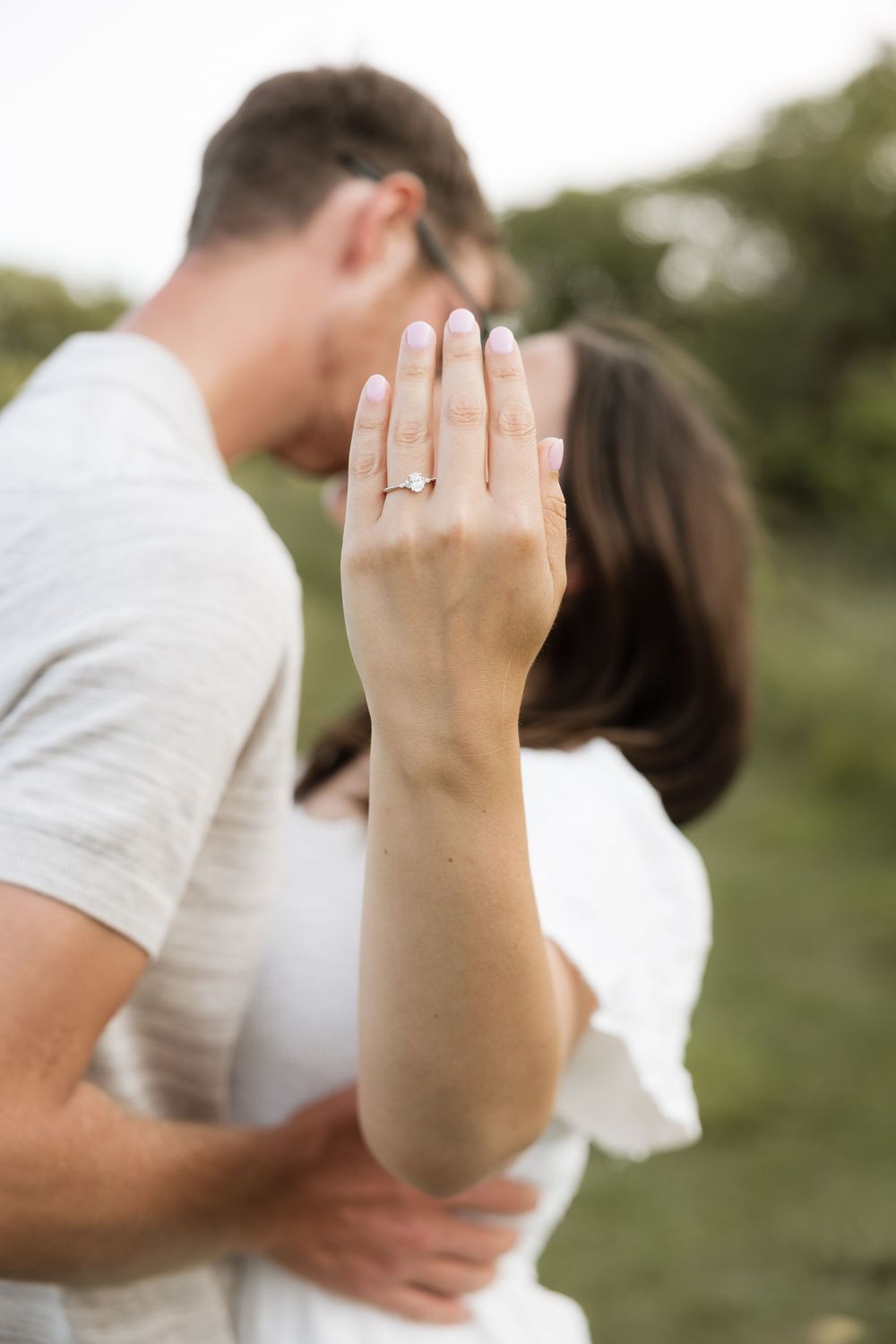 Couple kissing outdoors while woman shows off her engagement ring in the foreground.