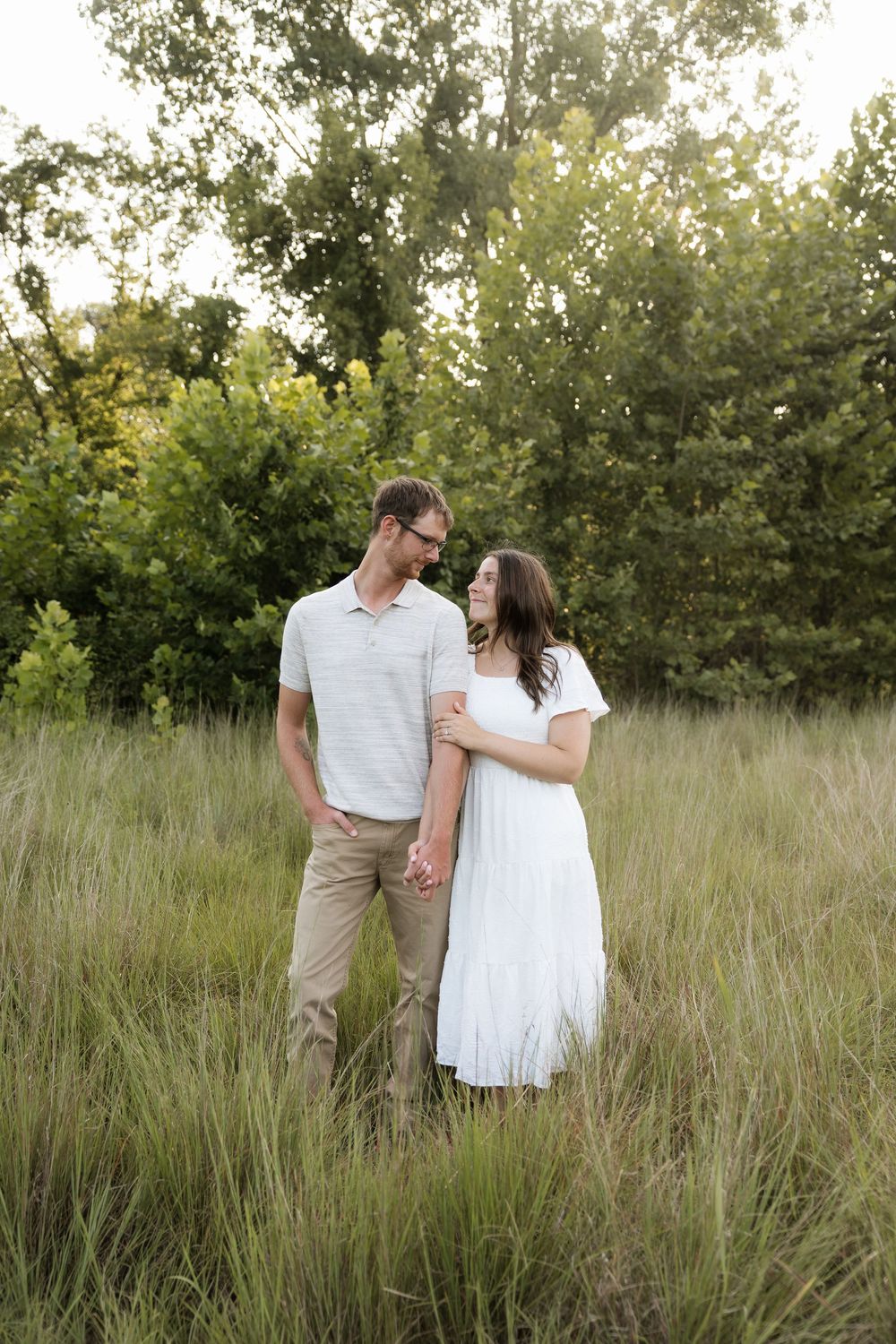 Couple holding hands and smiling at each other in a lush green field during golden hour.
