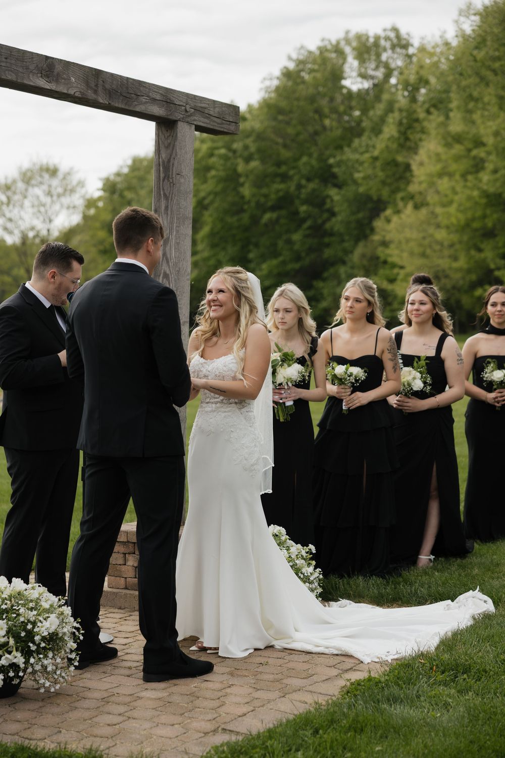 Bride smiling at groom during outdoor wedding ceremony with bridesmaids in black dresses standing beside wooden arbor.