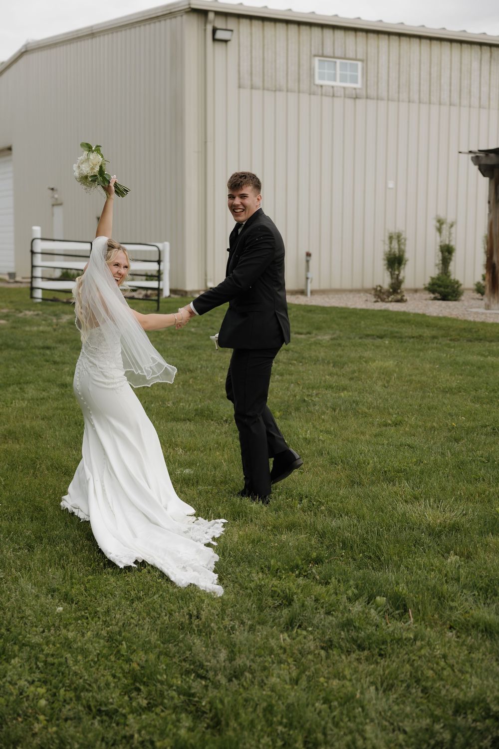 Joyful bride in white gown holding bouquet aloft while groom in black suit smiles, dancing on green lawn near barn.