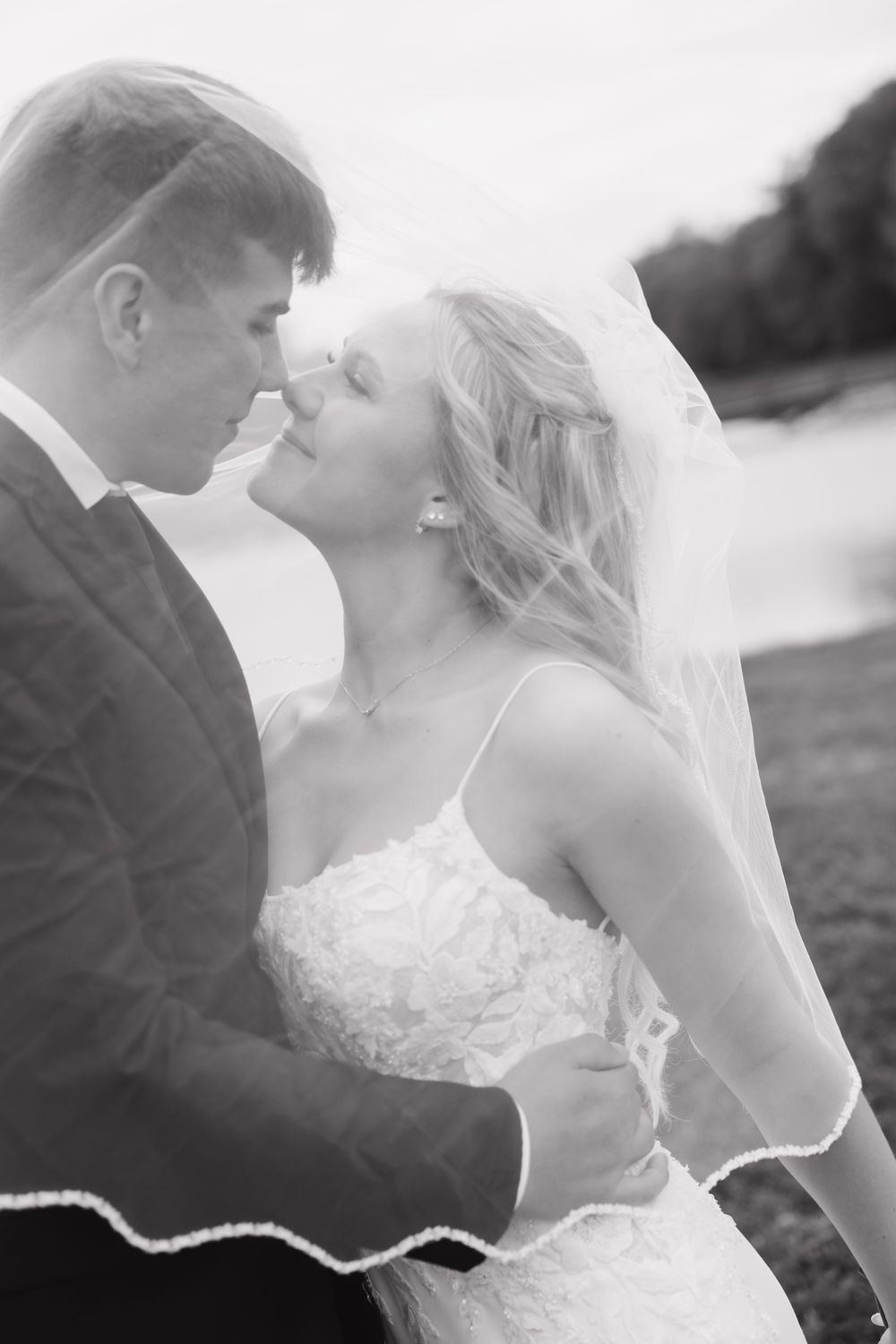 Black and white photo of a bride and groom sharing a romantic moment under a veil near a lake.