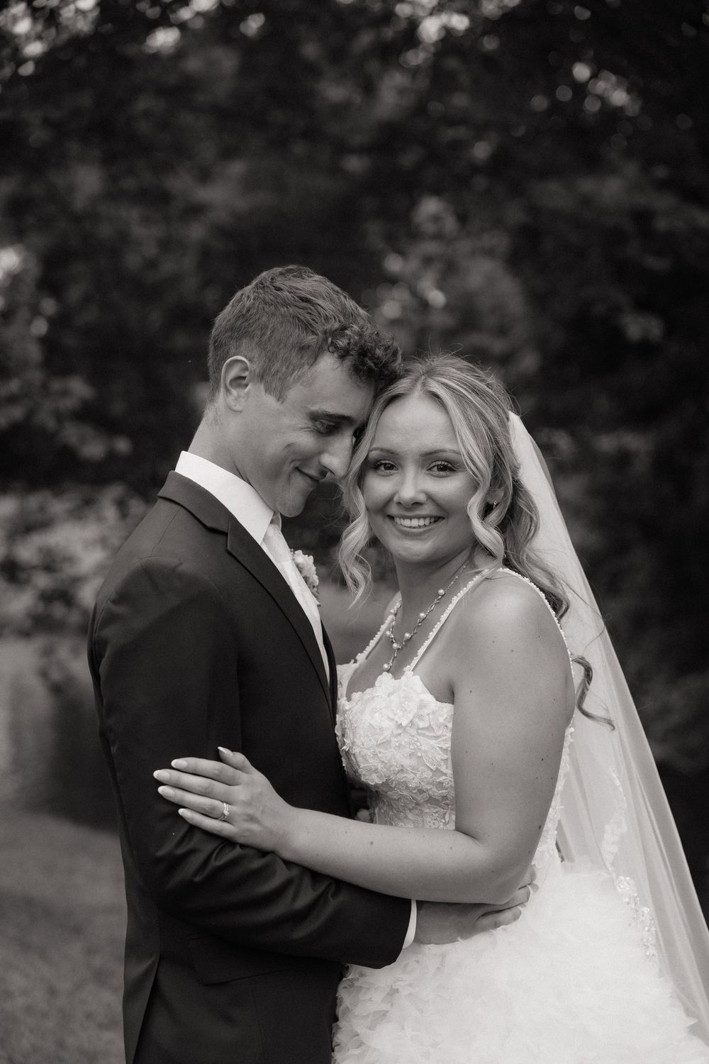 Black and white wedding portrait of smiling bride in lace gown and veil embracing groom in dark suit outdoors.