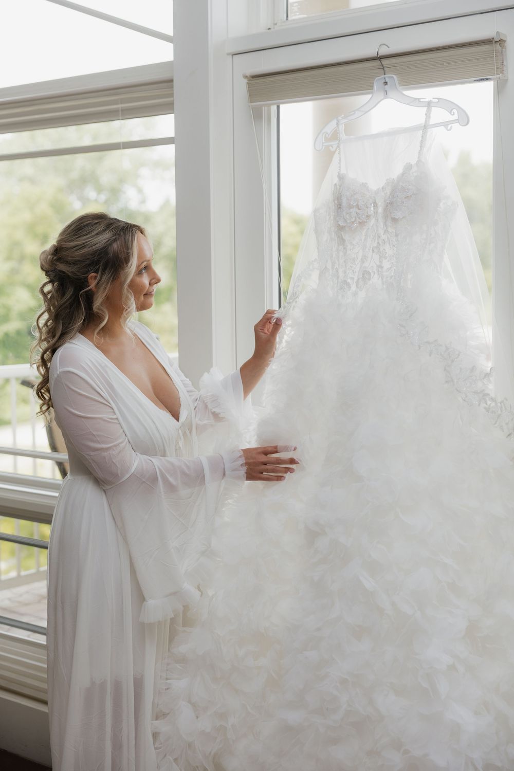 Bride in white robe admiring her lace wedding gown hanging by a bright window on her wedding day.