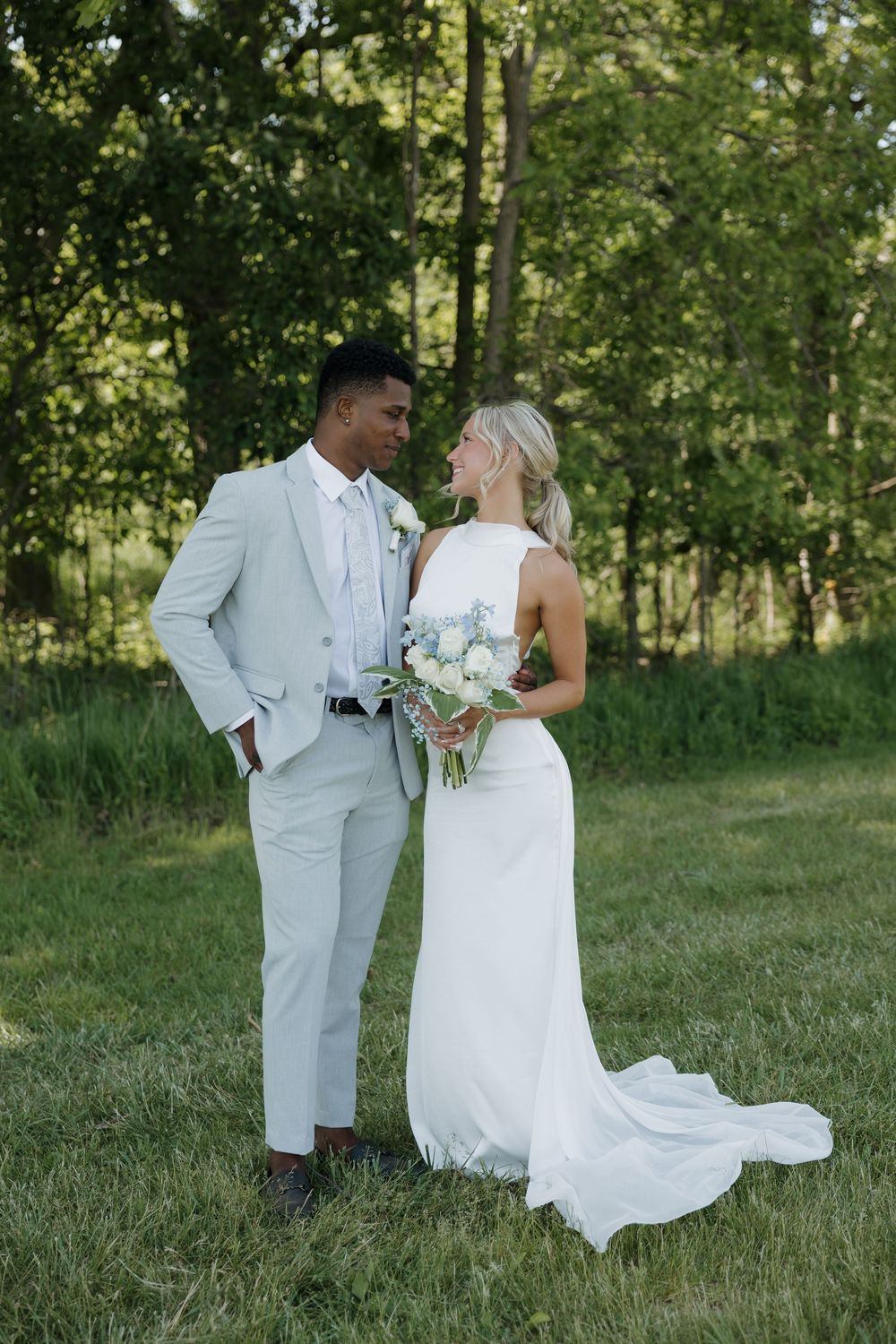 Interracial couple in wedding attire share a romantic moment outdoors, bride holding white floral bouquet.
