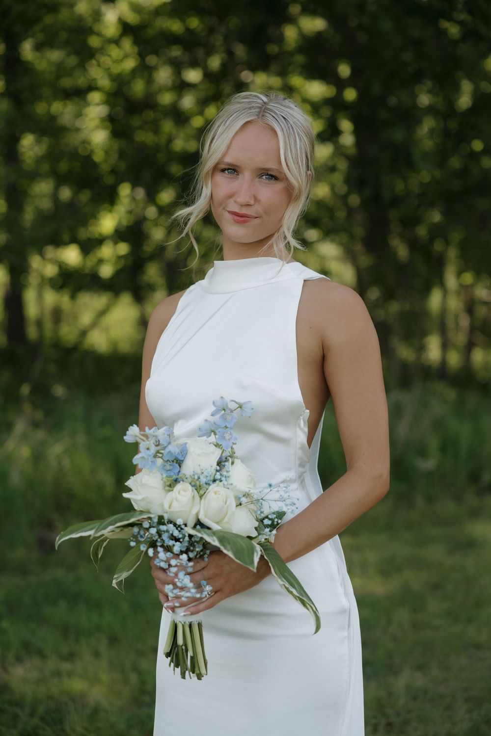Blonde bride in white halter gown holds blue and white floral bouquet outdoors with lush green trees in background.