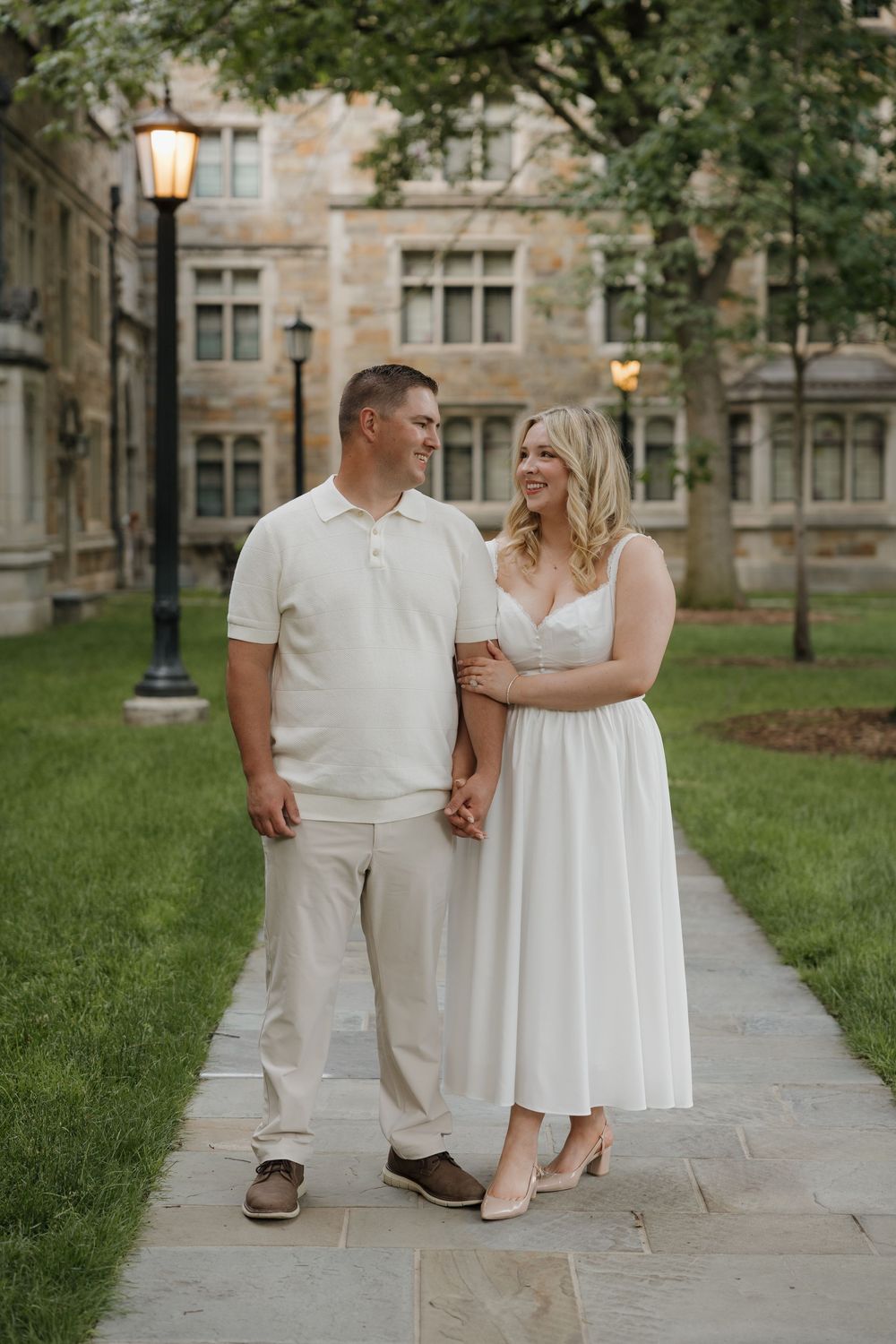 Couple in coordinating white and cream outfits holding hands on a stone pathway outside a classic brick building.