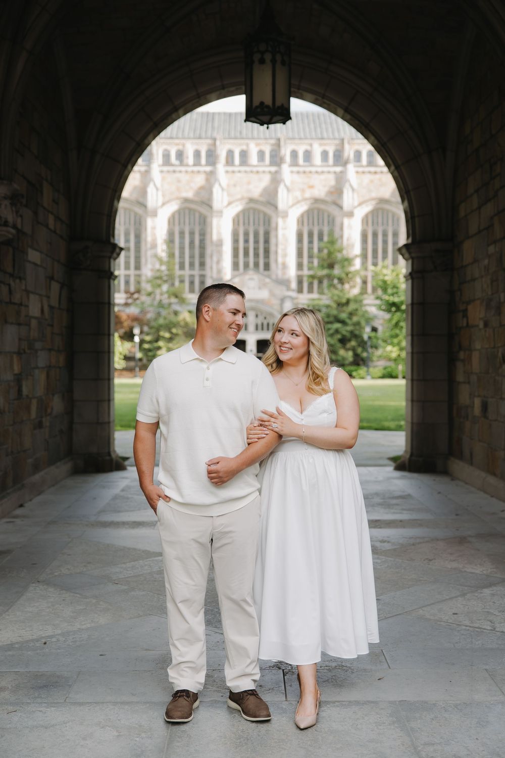 Couple in cream outfits smiling under stone arch with gothic university building in background.