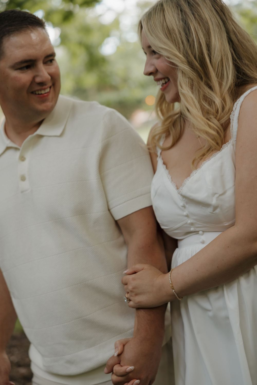 Smiling couple in white outfits holding hands outdoors, woman wearing engagement ring, man in polo shirt.
