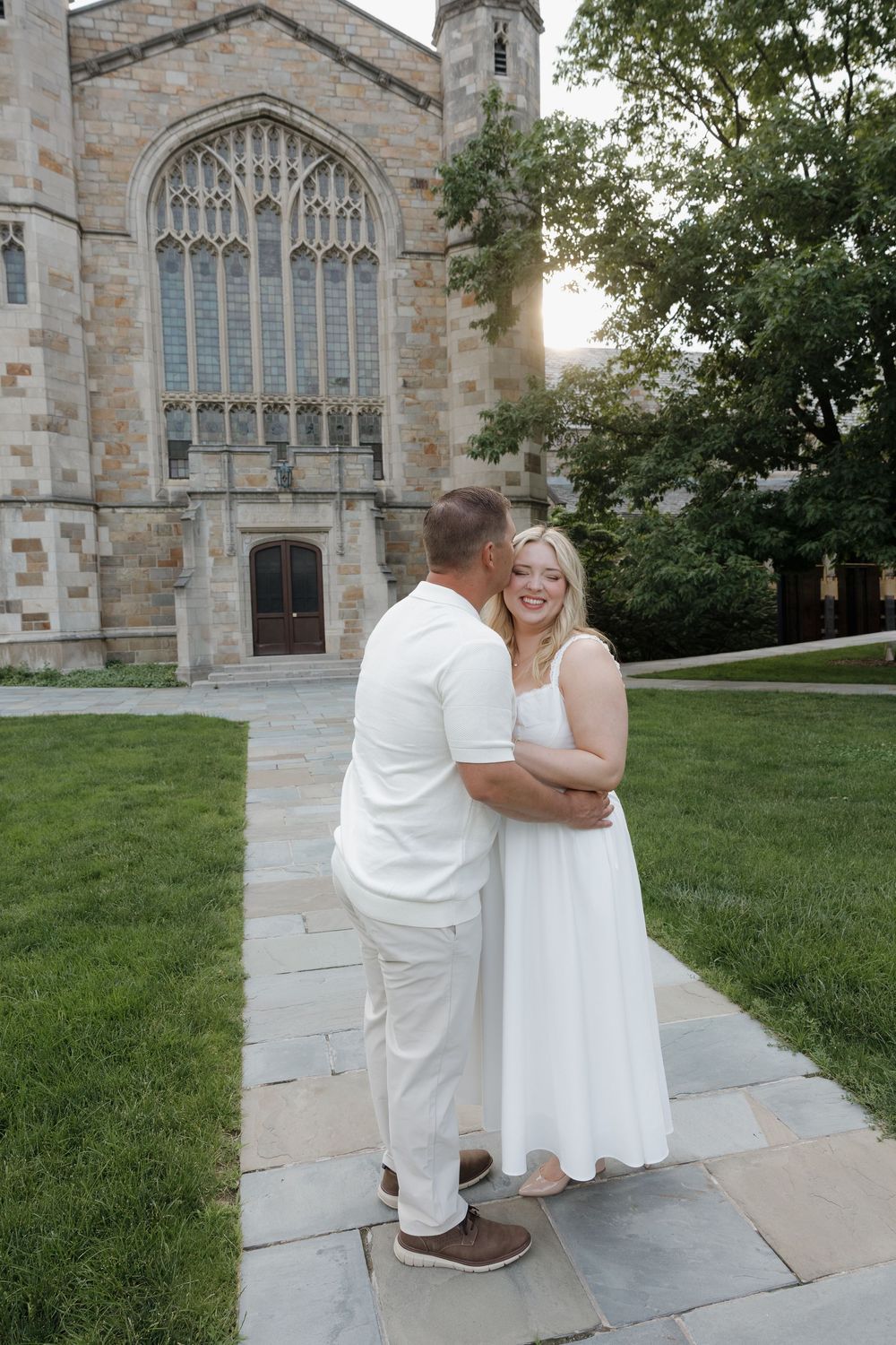 Couple in white outfits sharing a romantic moment on stone path before gothic stone church with large stained glass window.