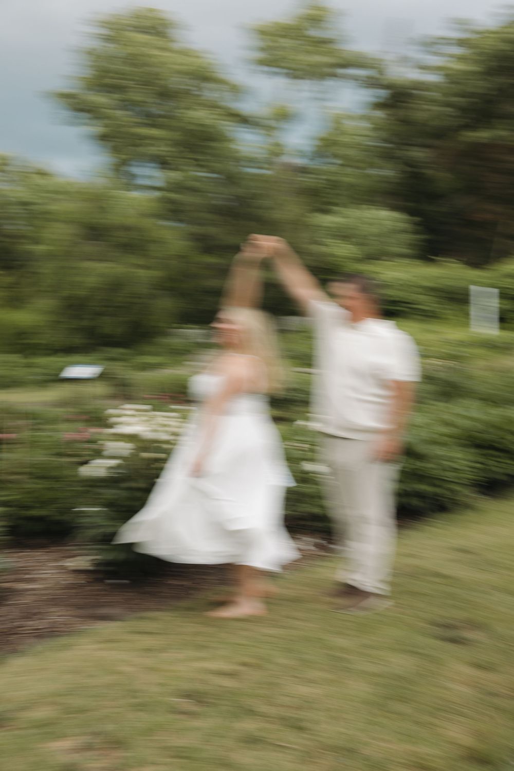 A couple dances outdoors in a lush garden, woman in white dress twirling, captured with artistic motion blur.