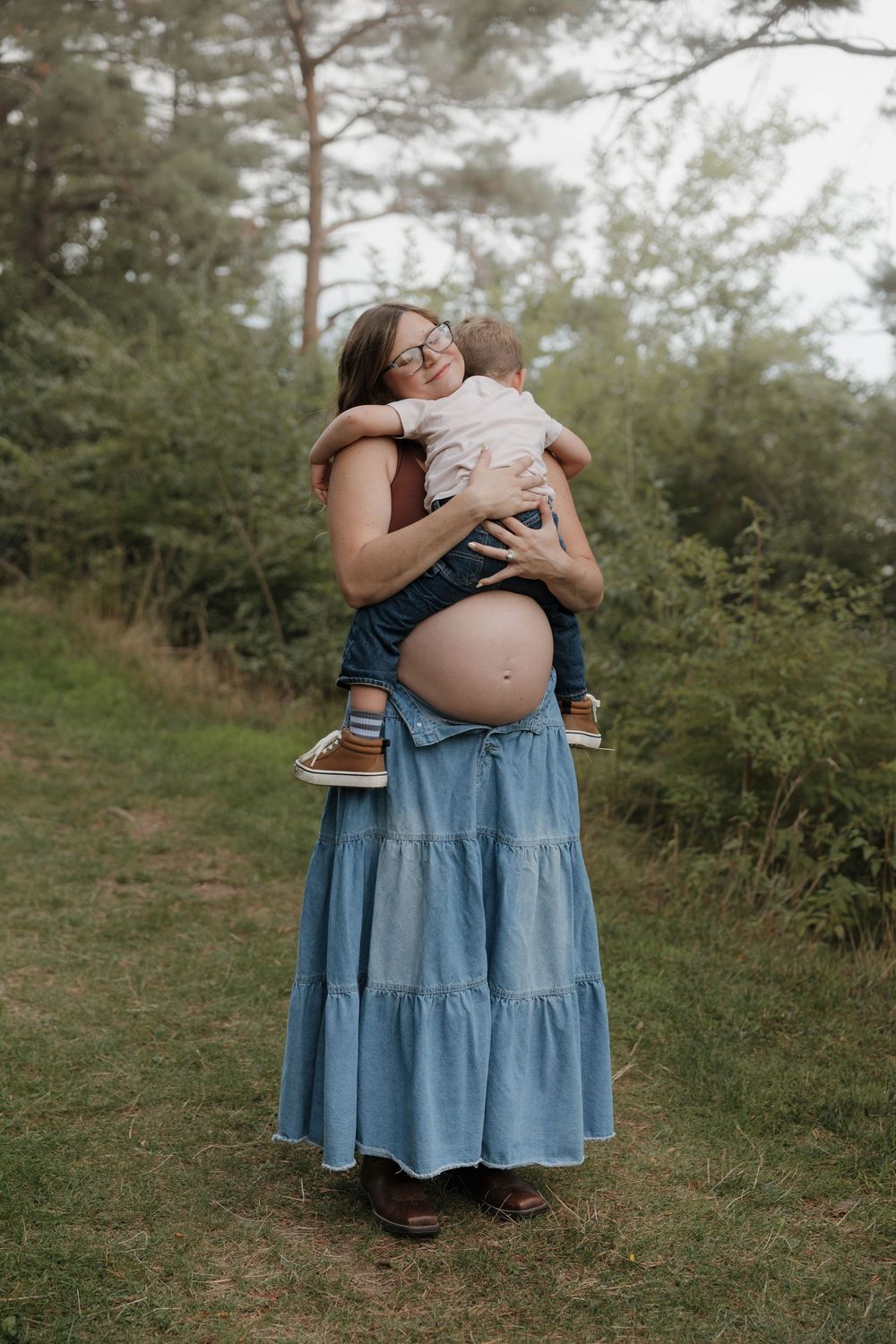 Pregnant woman in denim skirt hugging toddler outdoors in forest setting, showing baby bump.