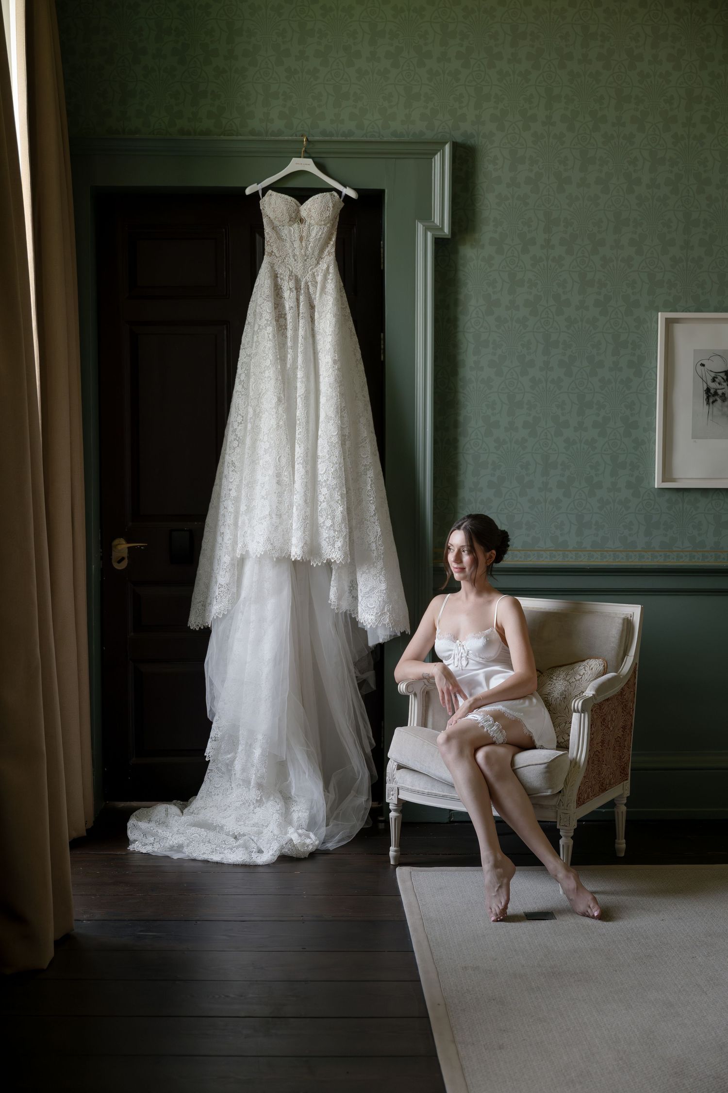 Beautiful lace wedding dress hanging in room with white armchair and sage green wallpapered walls.