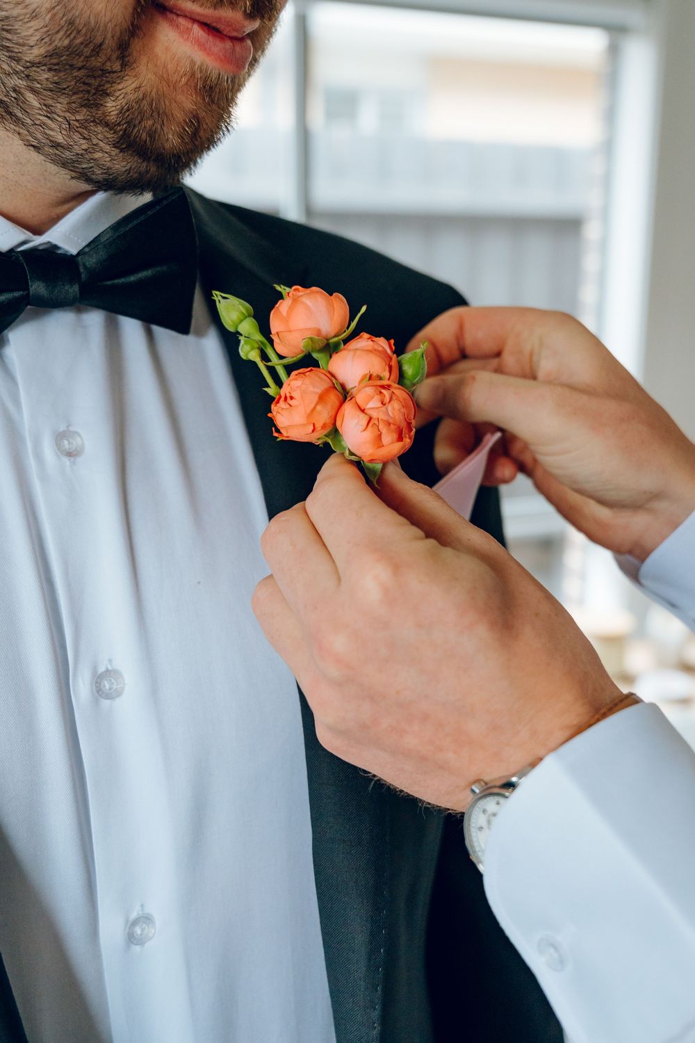 Close-up of hands pinning a coral boutonniere to a black tuxedo lapel.