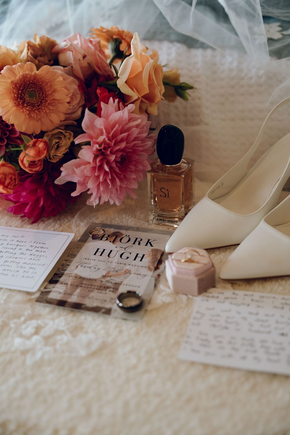 Wedding details with peach flowers, white heels, perfume bottle and invitation on lace.