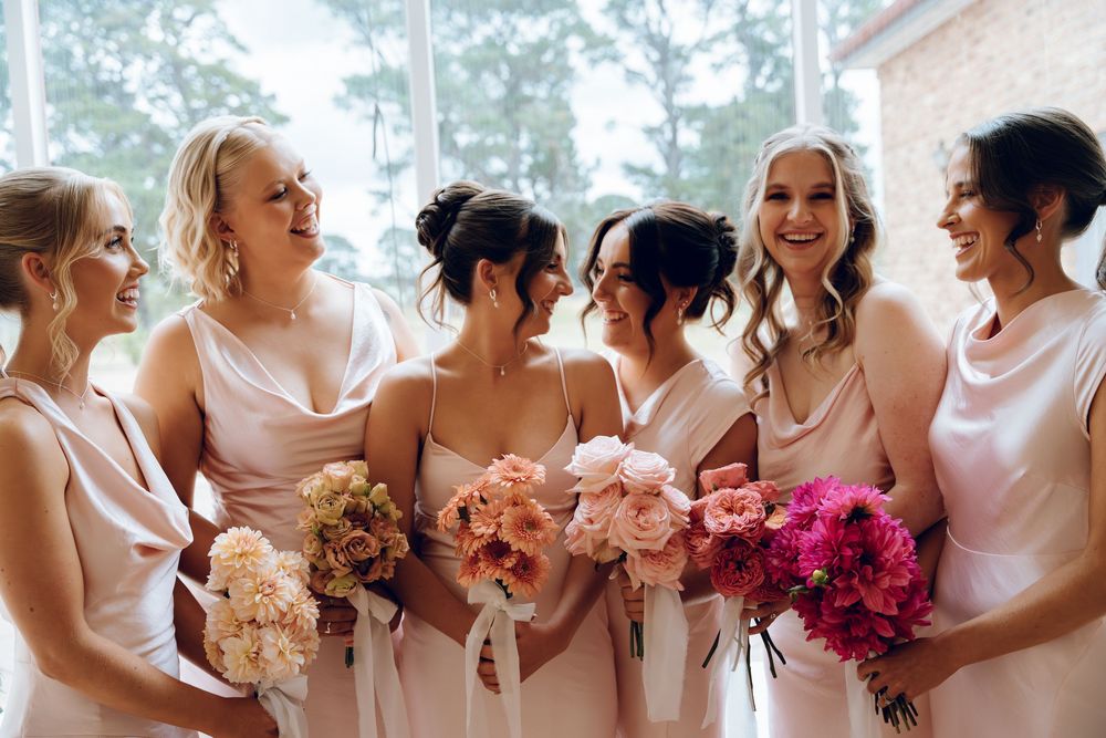 Bridesmaids in blush pink dresses holding colorful bouquets share a joyful moment.