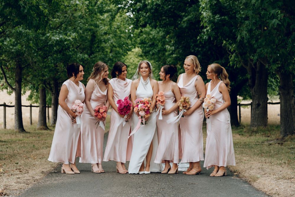 Bridesmaids in blush pink dresses pose with bouquets along tree-lined path.