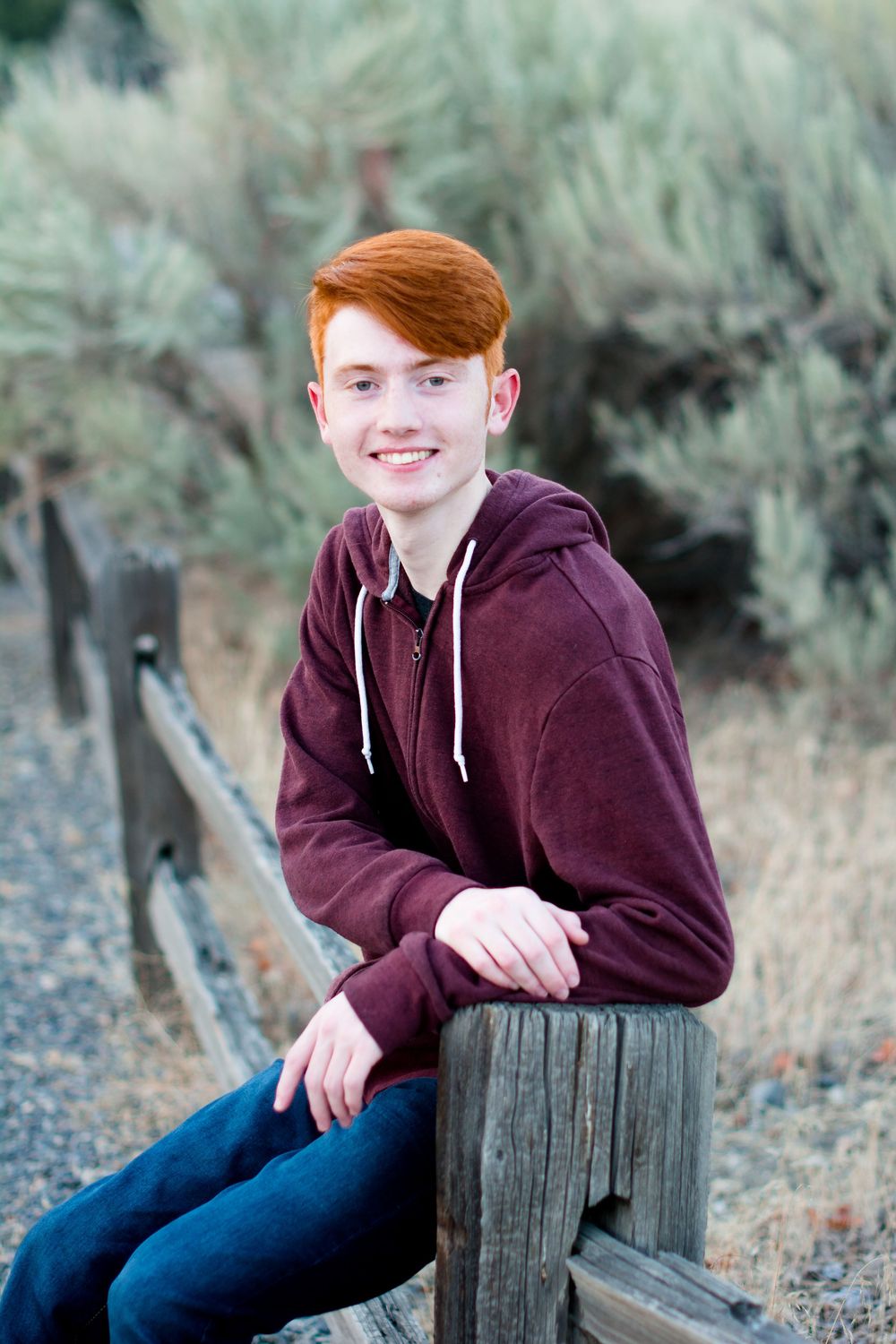 A student in a maroon hoodie and jeans sits casually outdoors on a wooden log against a blurred natural background.
