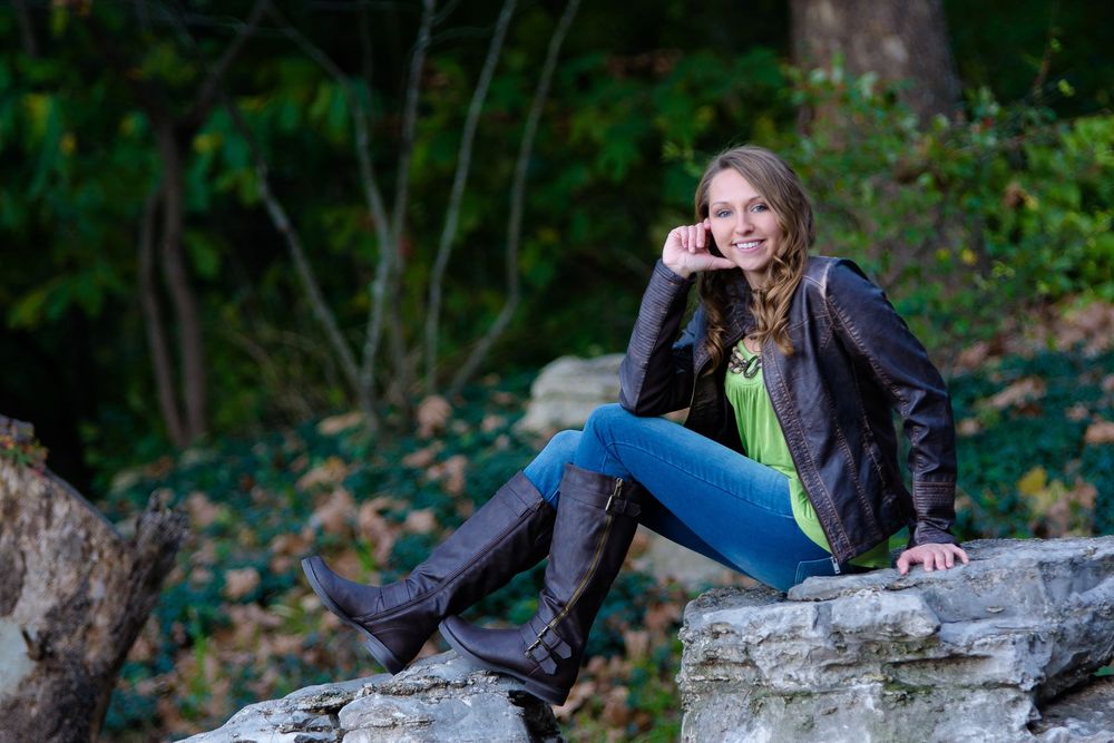 Person in leather jacket and jeans sits casually on rocks in autumn forest setting.