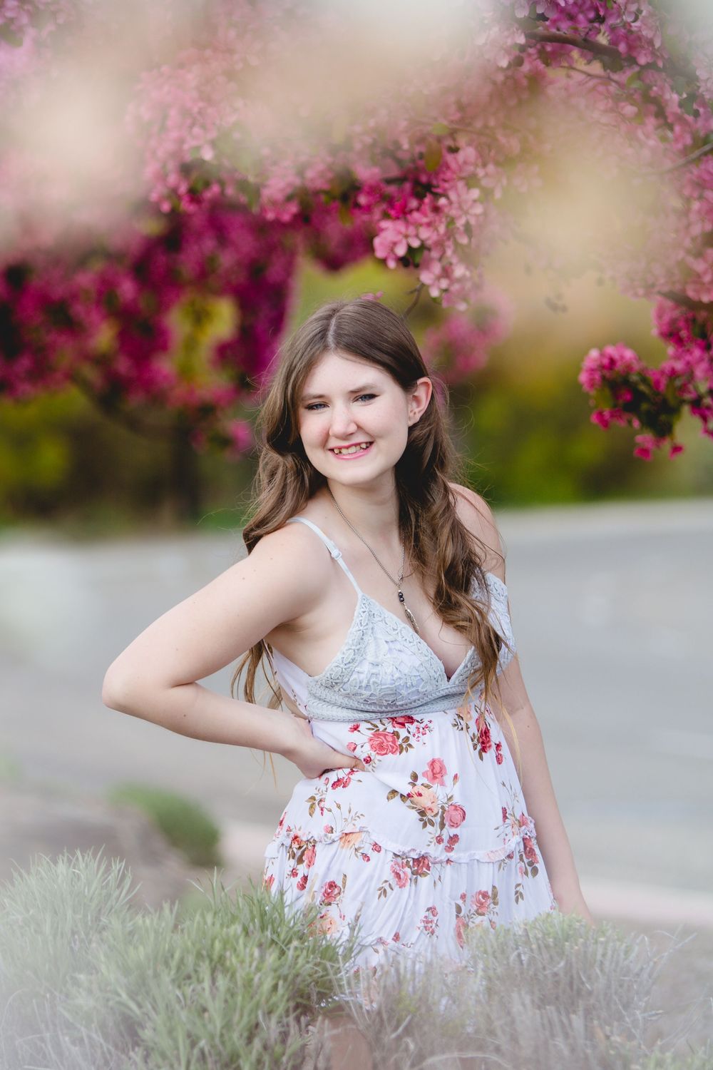 A person in a white and red floral dress poses playfully near blooming pink cherry blossoms on a spring day.