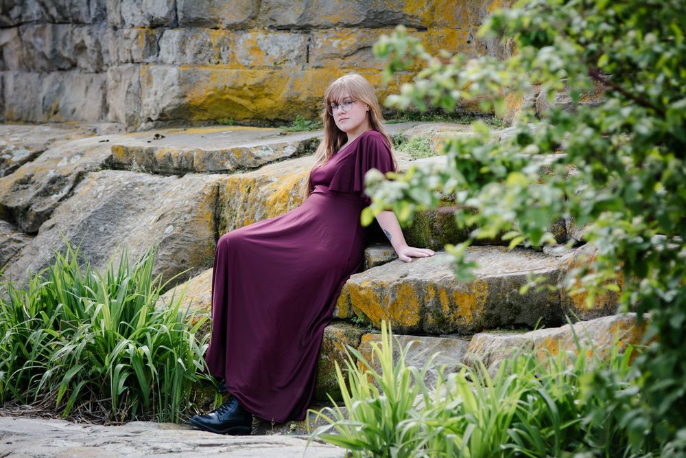 Woman in burgundy maxi dress sitting on stone steps surrounded by lush greenery in an outdoor garden setting.