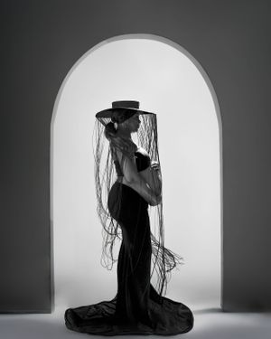 a black and white photograph of pregnant lady with a had standing between and arch in a black dress