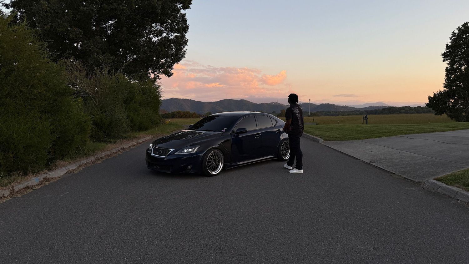 Person standing beside lowered dark blue Lexus sedan on empty road at dusk.