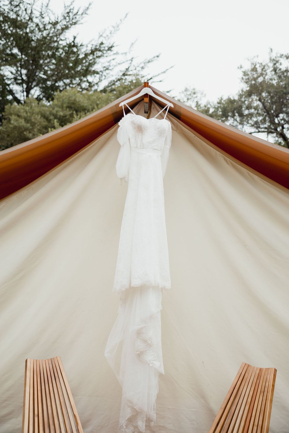 Elegant white wedding dress hanging from a canopy tent with wooden chairs below.