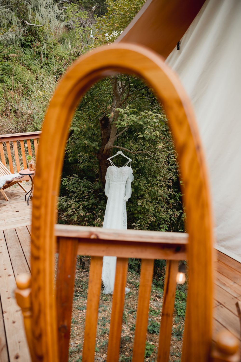 White wedding dress reflected in vintage mirror on wooden deck with tent backdrop.