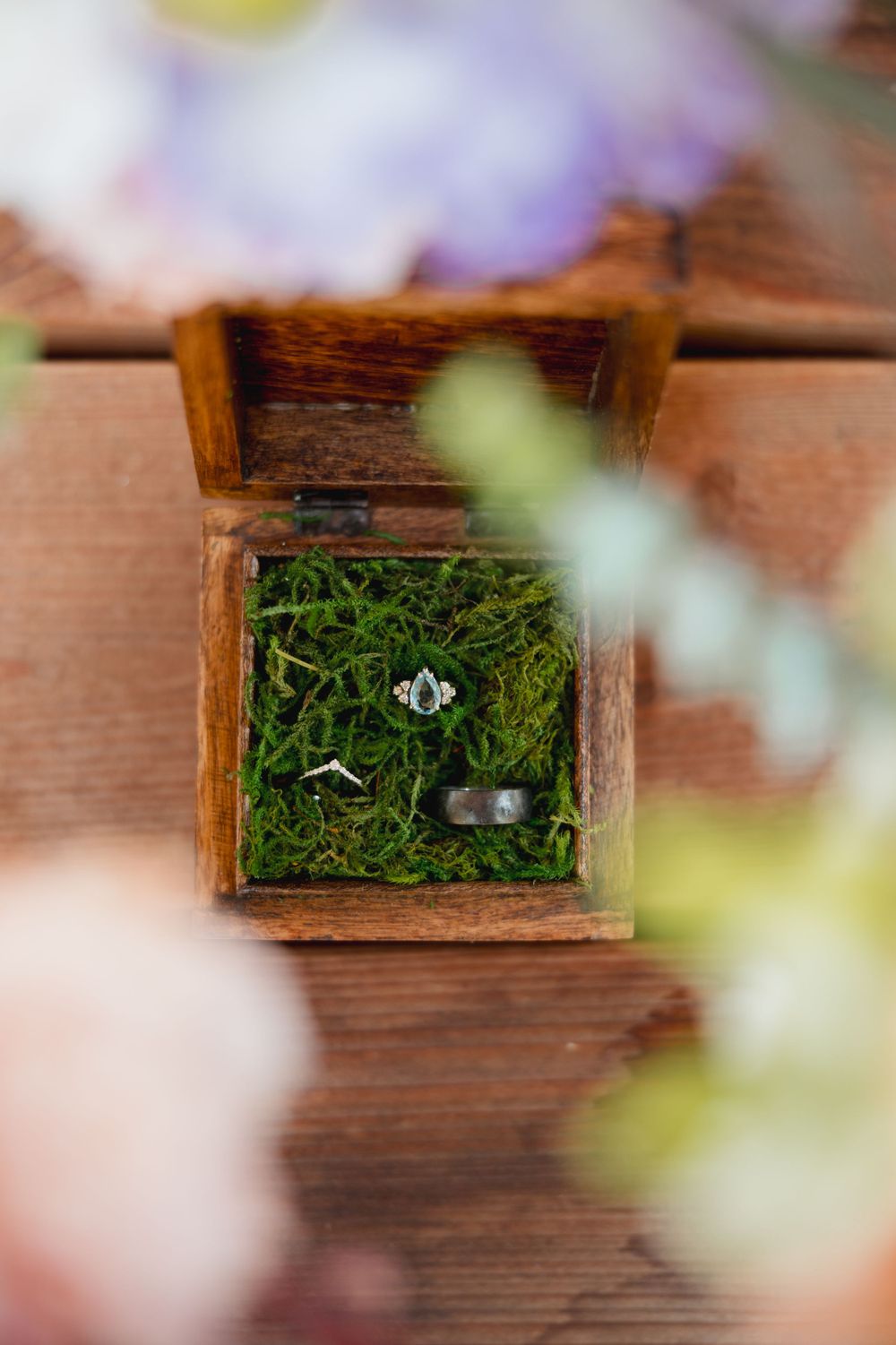 Wedding rings nestled in moss inside rustic wooden box with soft focus flowers.