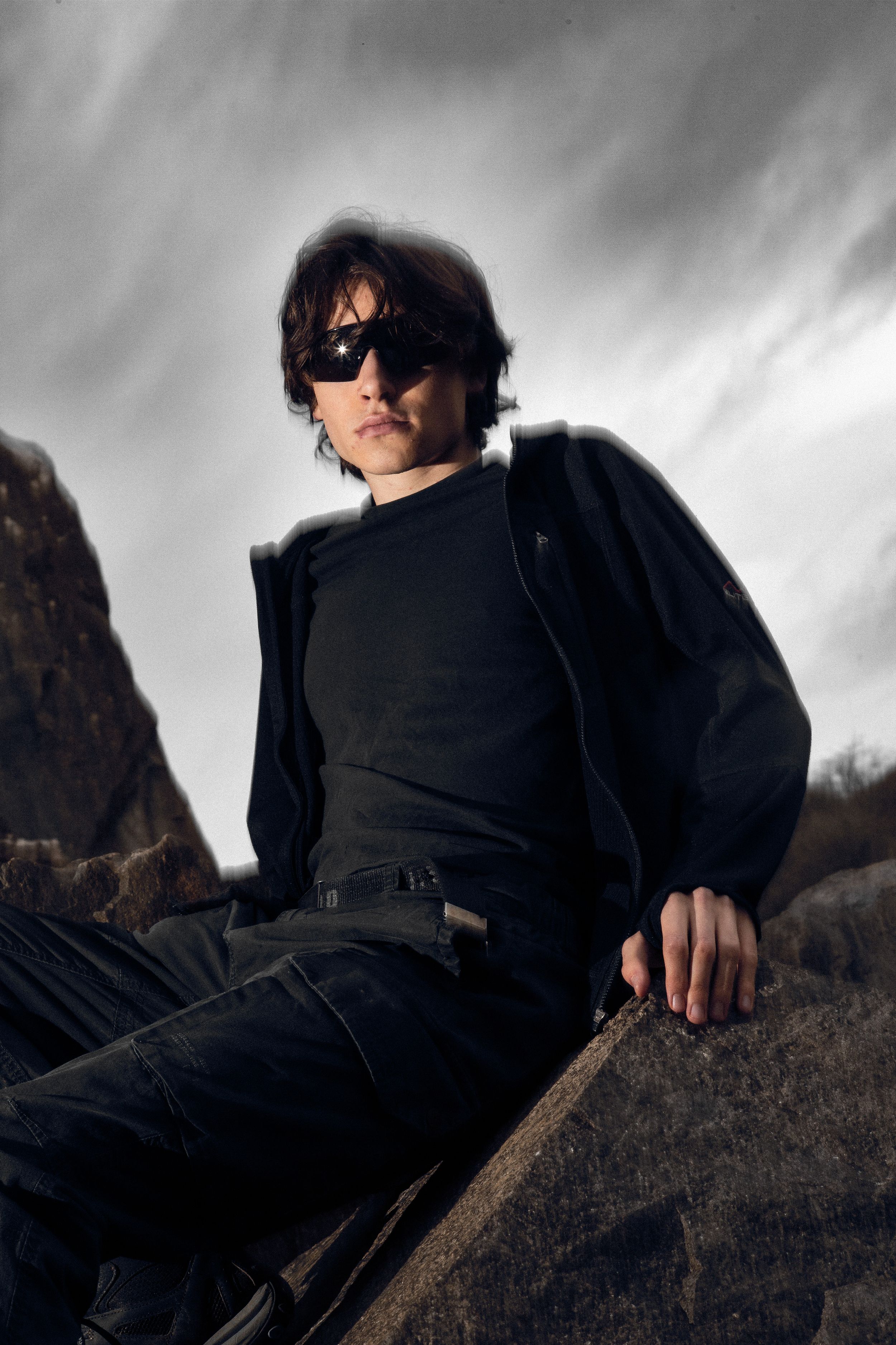 Figure in black clothing posing against dramatic cloudy sky with sunglasses.
