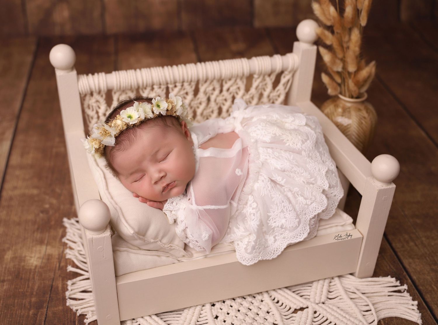 A sleeping newborn in lace dress on a miniature white wooden bed with floral crown.