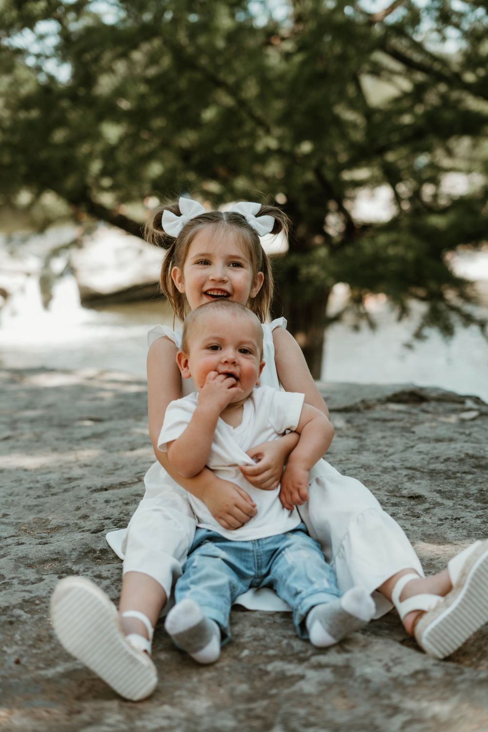 Two children in white clothing share a playful moment while sitting on rocks outdoors.