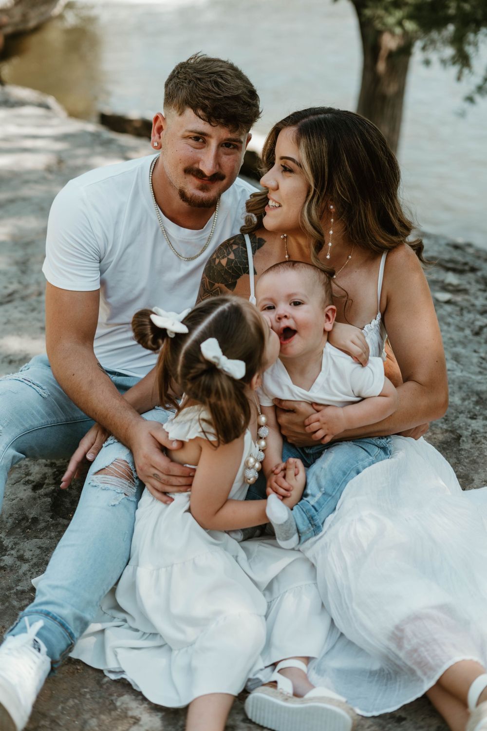 A family of four sits together on rocks by the water sharing a joyful moment.