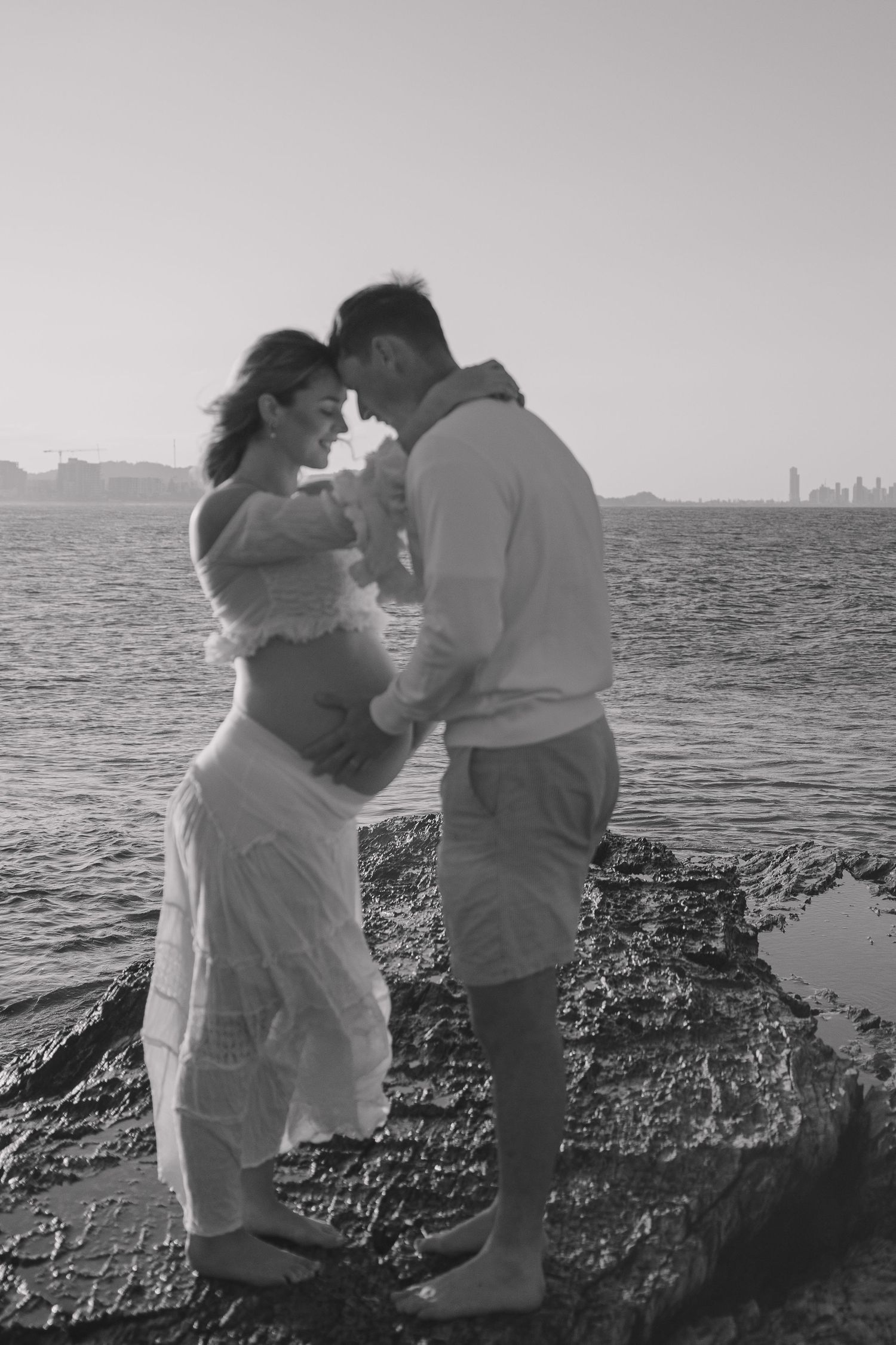 Black and white maternity photo of a couple touching foreheads on a rocky shore, her baby bump bare, with calm sea and skyline behind.