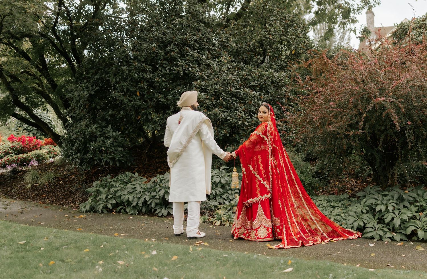 A wedding couple in traditional attire walking through a lush garden pathway.