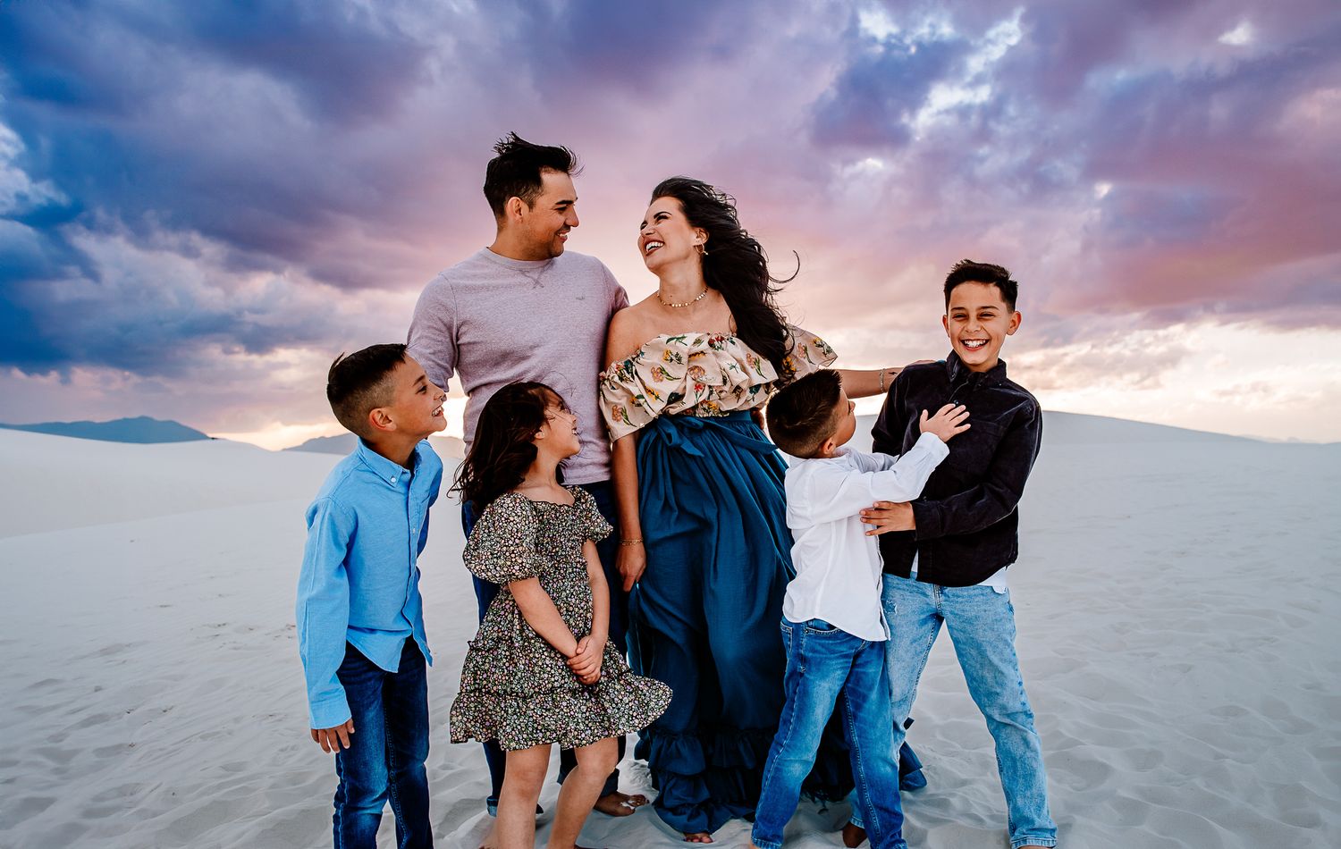 Family group poses together on scenic white sand dunes at sunset with dramatic purple clouds in background.