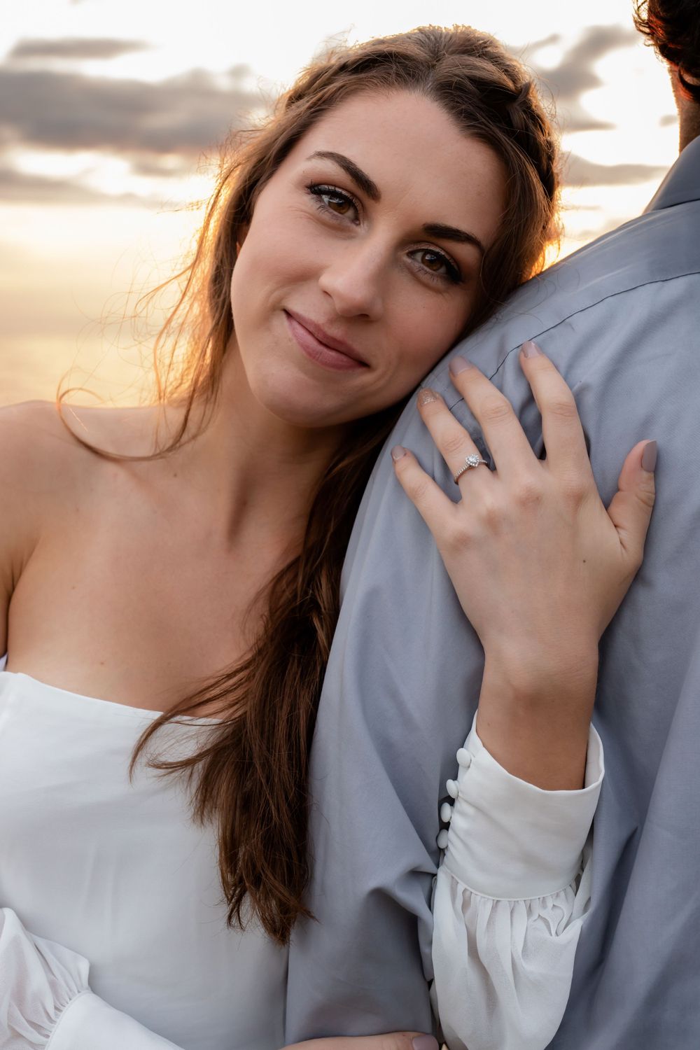 Bride wearing white dress smiles while embracing partner during sunset photoshoot.
