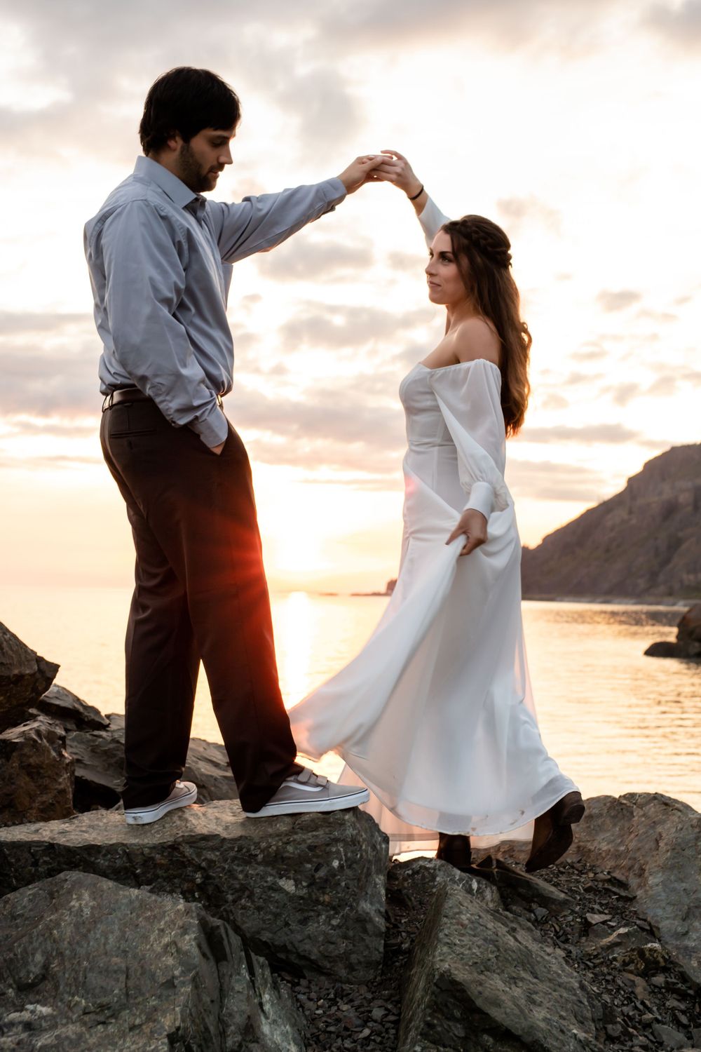 Couple in formal attire dance on rocky shore during golden hour sunset.