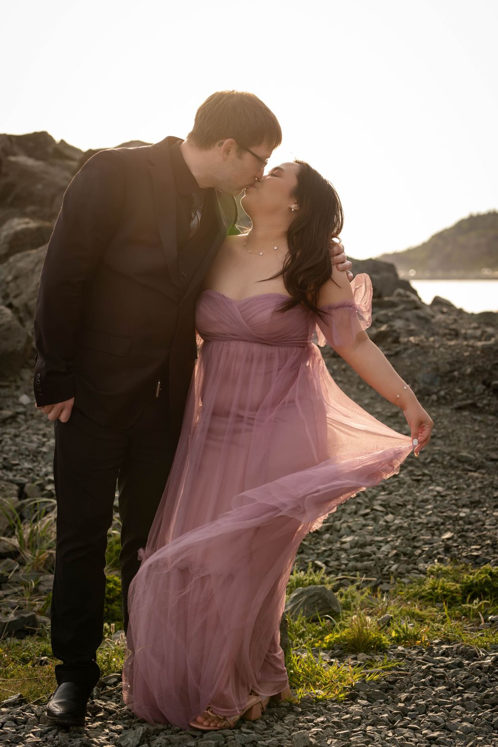 A romantic couple poses against a rocky coastal backdrop as the wind catches her flowing mauve dress at sunset.