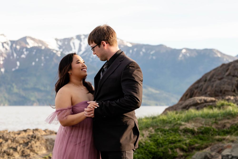 A couple shares an intimate moment on a cliffside overlooking snow-capped mountains and water.