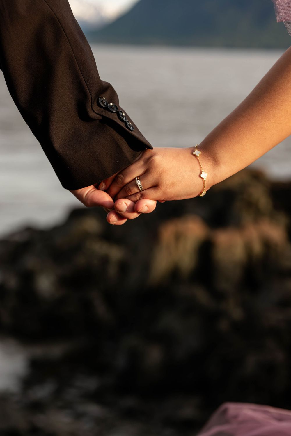 Close-up of two hands clasped together with a delicate bracelet against a rocky coastal background.