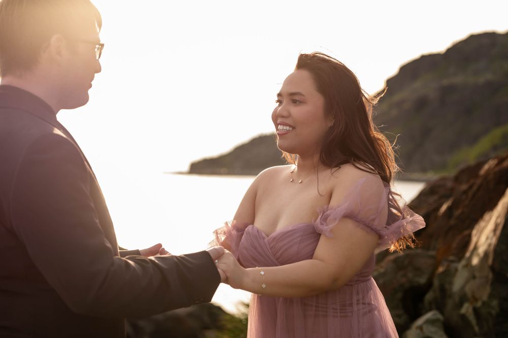 A couple embraces on a rocky cliff at golden hour with ocean views in the background.